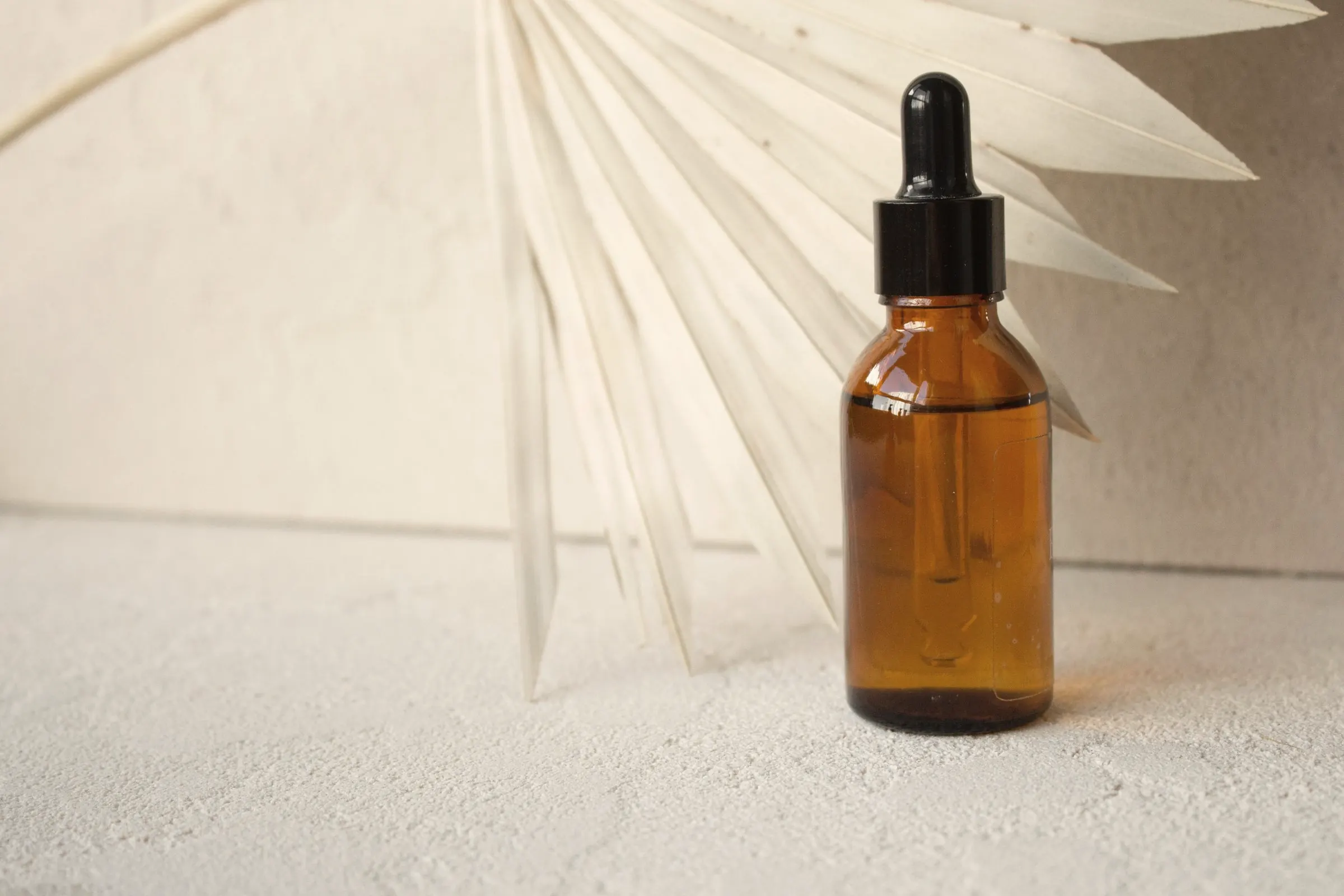 Amber glass dropper bottle beside a dried palm leaf on a plaster surface